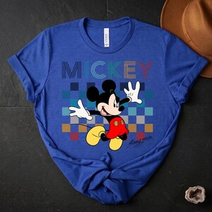 May include: Blue t-shirt featuring a graphic of Mickey Mouse with the word "MICKEY" in colorful block letters. Mickey is depicted with his arms outstretched, wearing red shorts, yellow shoes, and white gloves. The shirt has a crew neck and short sleeves.