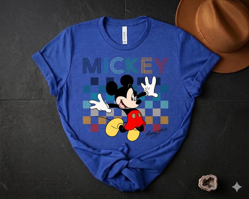 May include: Blue t-shirt featuring a graphic of Mickey Mouse with the word "MICKEY" in colorful block letters. Mickey is depicted with his arms outstretched, wearing red shorts, yellow shoes, and white gloves. The shirt has a crew neck and short sleeves.