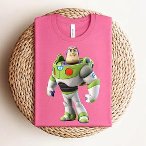 May include: A pink t-shirt featuring a graphic of Buzz Lightyear from Toy Story. The character is depicted in his signature green, white, and purple spacesuit. The shirt is folded and placed on a woven, circular surface.