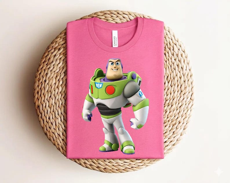 May include: A pink t-shirt featuring a graphic of Buzz Lightyear from Toy Story. The character is depicted in his signature green, white, and purple spacesuit. The shirt is folded and placed on a woven, circular surface.