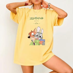 Może przedstawiać: Ż&oacute;łta koszulka z napisem "Zootopia" w kolorze zielonym, logo Disneya i słowem "Fan". Koszulka ma grafikę Nicka Wilde'a i Judy Hopps. Koszulka ma kr&oacute;tkie rękawy.