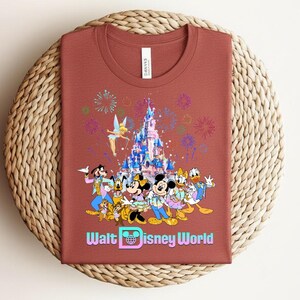 Może przedstawiać: Rdzawa koszulka z kolorową grafiką postaci Disneya, zamku, fajerwerk&oacute;w i napisem "Walt Disney World". Koszulka jest złożona na tkanej macie.