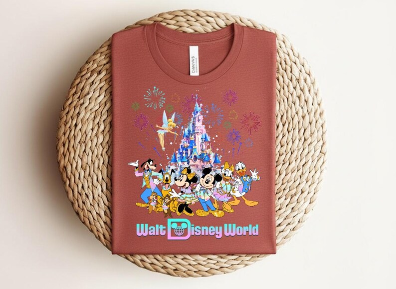 Może przedstawiać: Rdzawa koszulka z kolorową grafiką postaci Disneya, zamku, fajerwerk&oacute;w i napisem "Walt Disney World". Koszulka jest złożona na tkanej macie.