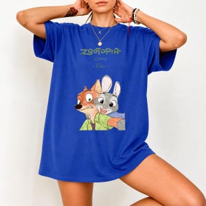 Może przedstawiać: Kr&oacute;lewsko niebieska koszulka z napisem "ZOOTOPIA" w kolorze zielonym i logo Disneya. Koszulka ma ilustrację Nicka Wilde'a i Judy Hopps robiących sobie selfie. Koszulka jest w stylu casual.