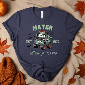 Może przedstawiać: Granatowa koszulka z motywem Mater z filmu Auta Disneya w tematyce Halloween. Projekt zawiera napis "MATER EST 1977 Disney Cars" z nietoperzami i pajęczyną. Grafika przedstawia Matera jako samoch&oacute;d zombie z zielonymi i pomarańczowymi akcentami.