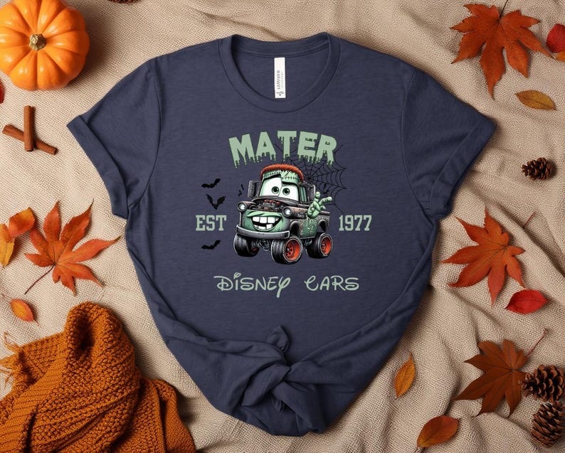 Może przedstawiać: Granatowa koszulka z motywem Mater z filmu Auta Disneya w tematyce Halloween. Projekt zawiera napis "MATER EST 1977 Disney Cars" z nietoperzami i pajęczyną. Grafika przedstawia Matera jako samoch&oacute;d zombie z zielonymi i pomarańczowymi akcentami.