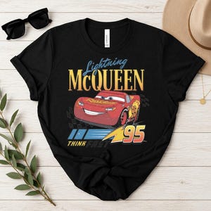 May include: Black t-shirt with a graphic of Lightning McQueen from the Cars film. The design includes the text "Lightning McQueen" in yellow and blue, the number "95", and the phrase "Think Fast".