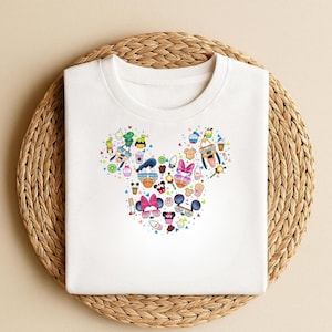 May include: White t-shirt with a Mickey Mouse head design made up of cartoon food, drinks, sunglasses, and character faces. The shirt is displayed on a woven, circular placemat.