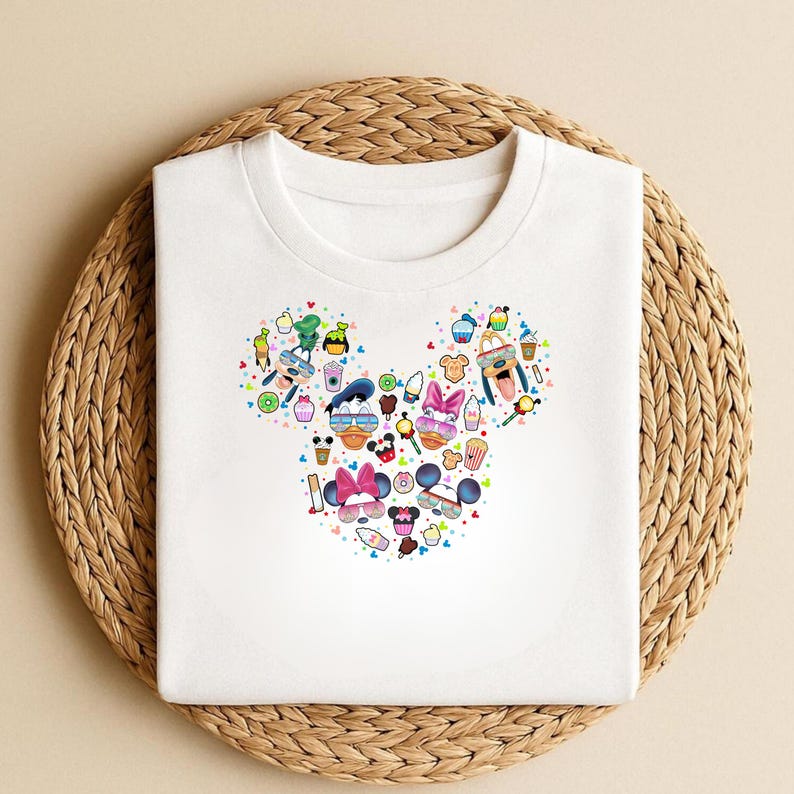 May include: White t-shirt with a Mickey Mouse head design made up of cartoon food, drinks, sunglasses, and character faces. The shirt is displayed on a woven, circular placemat.