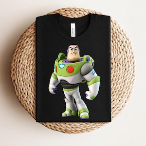 May include: Black t-shirt featuring a colorful graphic of Buzz Lightyear from Toy Story. The character is depicted in his signature green, white, and purple spacesuit. The shirt is displayed on a woven, circular surface.