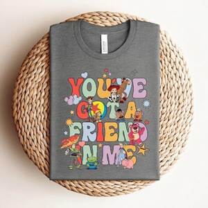 May include: A grey t-shirt with the text "You've Got a Friend in Me" in colourful, retro lettering. The design incorporates cartoon characters. The shirt is presented on a woven, circular surface.