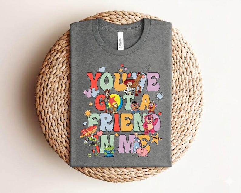 May include: A grey t-shirt with the text "You've Got a Friend in Me" in colourful, retro lettering. The design incorporates cartoon characters. The shirt is presented on a woven, circular surface.