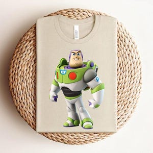 May include: A beige t-shirt featuring a full-color graphic of Buzz Lightyear from Toy Story. The character is depicted in his signature space ranger suit, with green, white, purple, and black accents. The shirt is folded on a woven, circular surface.