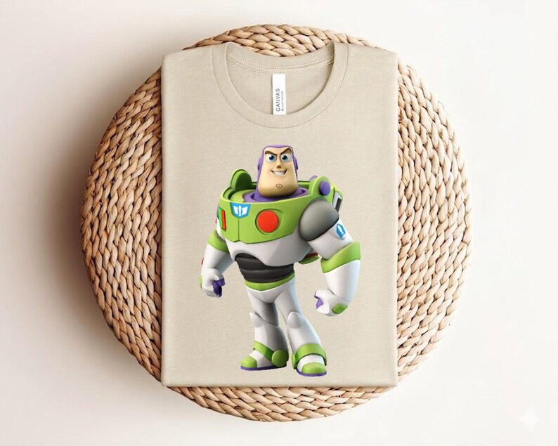 May include: A beige t-shirt featuring a full-color graphic of Buzz Lightyear from Toy Story. The character is depicted in his signature space ranger suit, with green, white, purple, and black accents. The shirt is folded on a woven, circular surface.
