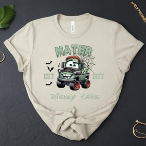 Może przedstawiać: Beżowa koszulka z grafiką Mater z filmu Auta Disneya w stylu zombie. Projekt zawiera napisy "MATER", "EST 1977" i "Disney Cars" w kolorze zielonym, z nietoperzami i pajęczyną.