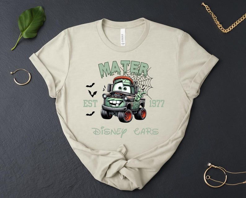 Może przedstawiać: Beżowa koszulka z grafiką Mater z filmu Auta Disneya w stylu zombie. Projekt zawiera napisy "MATER", "EST 1977" i "Disney Cars" w kolorze zielonym, z nietoperzami i pajęczyną.