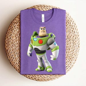 May include: A purple t-shirt featuring a graphic of Buzz Lightyear from Toy Story. The character is depicted in his signature green, white, and purple spacesuit. The shirt is displayed on a woven, circular placemat.