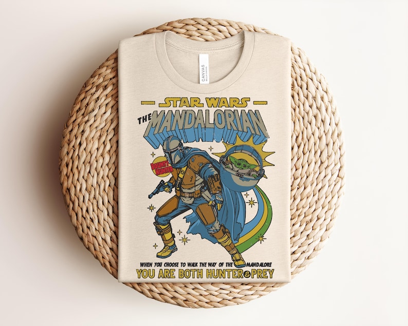 K&ouml;nnte beinhalten: Beiges T-Shirt mit einer Grafik von Star Wars The Mandalorian. Das Design zeigt die Mandalorian Figur mit Grogu und den Text "STAR WARS THE MANDALORIAN". Es enth&auml;lt auch das Zitat "WHEN YOU CHOOSE TO WALK THE WAY OF THE MANDALORE YOU ARE BOTH HUNTER & PREY."
