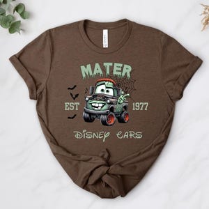 Może przedstawiać: Brązowa koszulka z kresk&oacute;wkowym wizerunkiem Złomka z filmu Auta Disneya w tematyce Halloween. Projekt zawiera napis "MATER EST 1977 DISNEY CARS" z nietoperzami i pajęczyną.