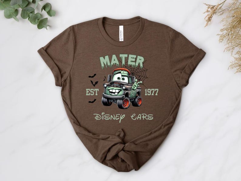 Może przedstawiać: Brązowa koszulka z kresk&oacute;wkowym wizerunkiem Złomka z filmu Auta Disneya w tematyce Halloween. Projekt zawiera napis "MATER EST 1977 DISNEY CARS" z nietoperzami i pajęczyną.