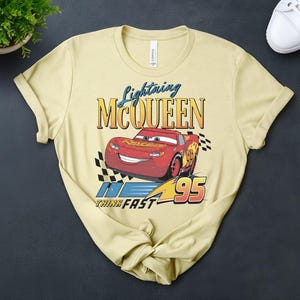 May include: A pale yellow t-shirt with a graphic of Lightning McQueen from the film Cars. The design includes the text "Lightning McQueen" above a red race car with the number 95, and the words "Think Fast".