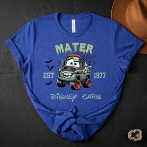 Może przedstawiać: Niebieska koszulka z motywem zombie Mater z filmu Disney Cars. Grafika zawiera napisy "MATER", "EST 1977" i "Disney Cars", a także nietoperze i pajęczynę. Koszulka jest wykonana z miękkiego materiału.