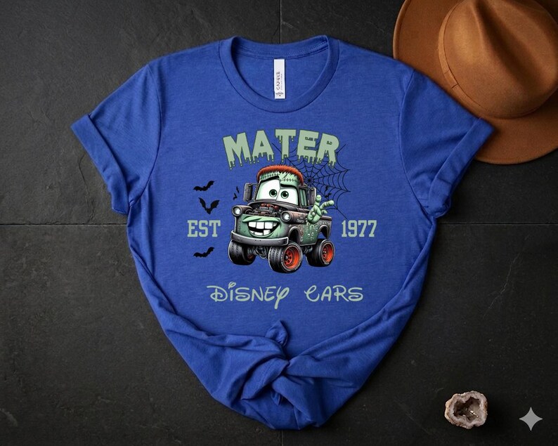Może przedstawiać: Niebieska koszulka z motywem zombie Mater z filmu Disney Cars. Grafika zawiera napisy "MATER", "EST 1977" i "Disney Cars", a także nietoperze i pajęczynę. Koszulka jest wykonana z miękkiego materiału.