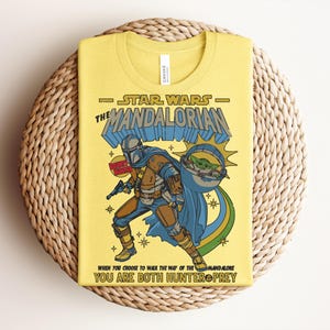 K&ouml;nnte beinhalten: Gelbes T-Shirt mit einer Grafik von The Mandalorian aus Star Wars. Das Design enth&auml;lt den Text "STAR WARS THE MANDALORIAN" und das Zitat "WHEN YOU CHOOSE TO WALK THE WAY OF THE MANDALORE YOU ARE BOTH HUNTER PREY". Die Illustration zeigt den Mandalorianer mit Grogu.