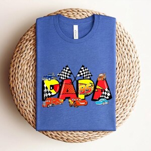 Puede incluir: Camiseta azul con la palabra "PAPA" en letras coloridas con tem&aacute;tica de coches. El dise&ntilde;o incluye banderas de carreras, el n&uacute;mero "95" y varios coches de dibujos animados. La camiseta est&aacute; doblada sobre una superficie circular tejida.