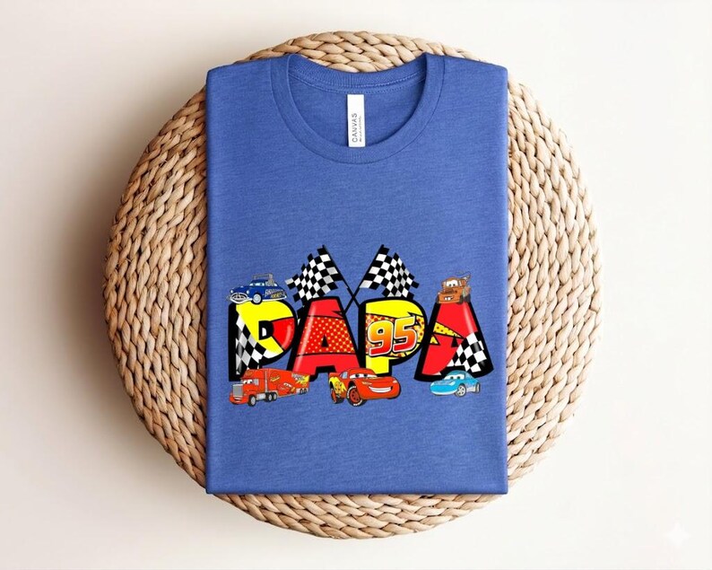 Puede incluir: Camiseta azul con la palabra "PAPA" en letras coloridas con tem&aacute;tica de coches. El dise&ntilde;o incluye banderas de carreras, el n&uacute;mero "95" y varios coches de dibujos animados. La camiseta est&aacute; doblada sobre una superficie circular tejida.