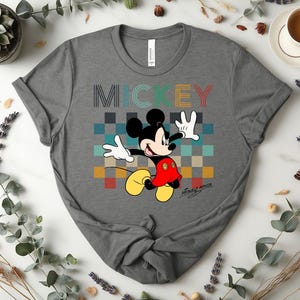 May include: Gray t-shirt featuring a colorful graphic of Mickey Mouse. The word "MICKEY" is printed above the character in a retro font. Mickey Mouse is depicted in his classic outfit, with a red shirt, yellow shoes, and white gloves. The shirt is made of a soft material.