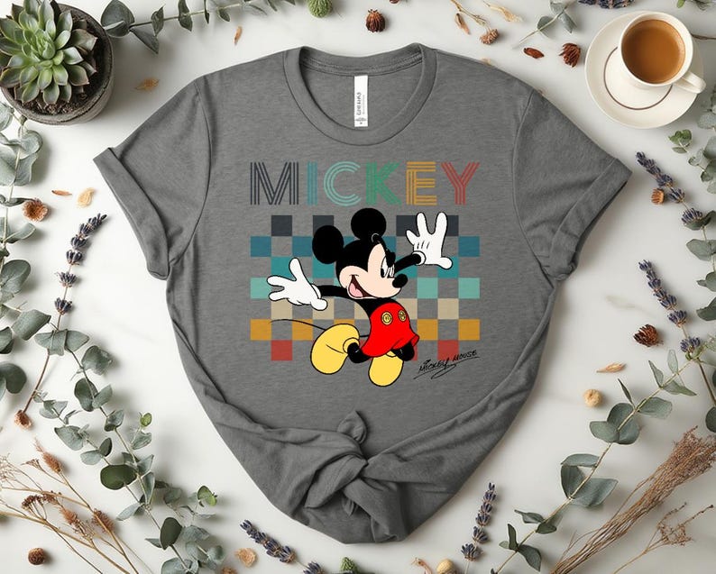 May include: Gray t-shirt featuring a colorful graphic of Mickey Mouse. The word "MICKEY" is printed above the character in a retro font. Mickey Mouse is depicted in his classic outfit, with a red shirt, yellow shoes, and white gloves. The shirt is made of a soft material.
