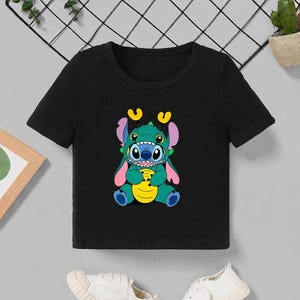 May include: Black crop top featuring a colorful graphic of a cartoon character with blue, green, yellow, and pink accents. The character has large eyes and is holding a yellow object. White sneakers are at the bottom.