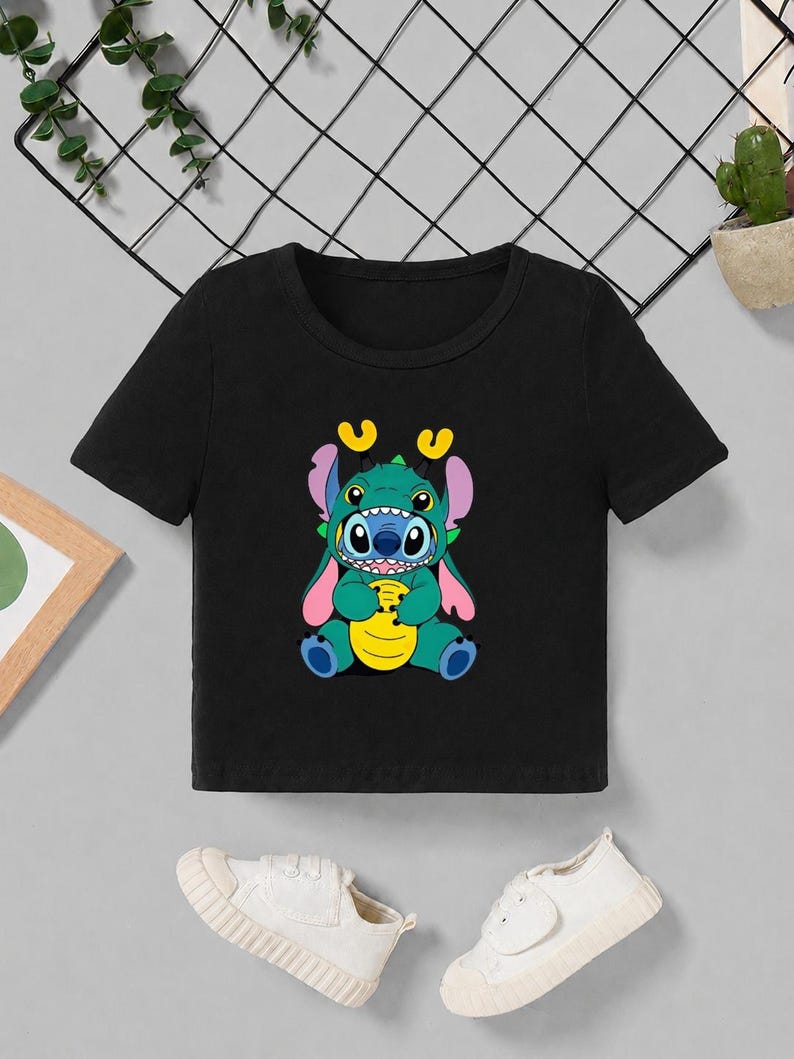 May include: Black crop top featuring a colorful graphic of a cartoon character with blue, green, yellow, and pink accents. The character has large eyes and is holding a yellow object. White sneakers are at the bottom.