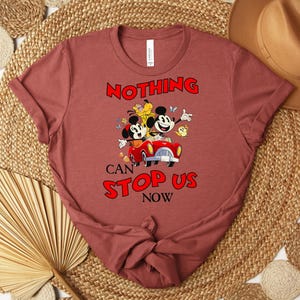 Puede incluir: Una camiseta granate con un dibujo animado de Mickey y Minnie Mouse en un coche rojo, con el texto "NOTHING CAN STOP US NOW" en rojo. El dise&ntilde;o incluye otros personajes de dibujos animados y mariposas. La camiseta est&aacute; doblada.