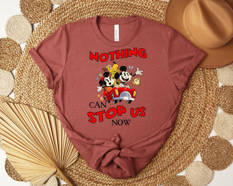 Puede incluir: Una camiseta granate con un dibujo animado de Mickey y Minnie Mouse en un coche rojo, con el texto "NOTHING CAN STOP US NOW" en rojo. El dise&ntilde;o incluye otros personajes de dibujos animados y mariposas. La camiseta est&aacute; doblada.