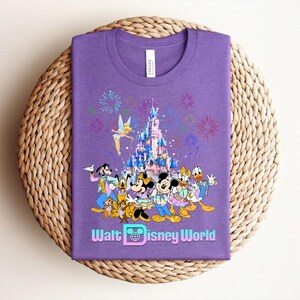 Może przedstawiać: Fioletowa koszulka z kolorową grafiką postaci Disneya, w tym Myszki Miki, Myszki Minnie, Kaczora Donalda i Goofy'ego, na tle zamku i fajerwerk&oacute;w. Tekst "Walt Disney World" jest wyświetlany na dole.