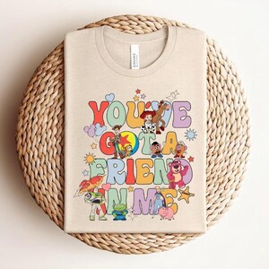 May include: Beige t-shirt with the text "You've Got a Friend in Me" in colourful, retro-style lettering. The shirt features cartoon characters from the Toy Story movie, including Woody, Buzz Lightyear, and others. The shirt is folded on a woven surface.