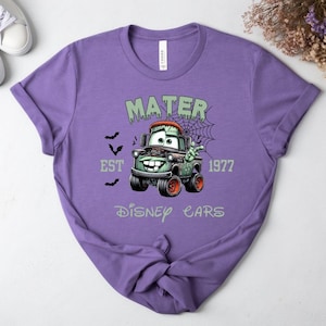 Może przedstawiać: Fioletowa koszulka z nadrukiem Mater z filmu Auta Disneya jako zombie. Projekt zawiera napisy "MATER", "EST 1977" i "Disney Cars", z nietoperzami i pajęczyną. Koszulka ma okrągły dekolt.