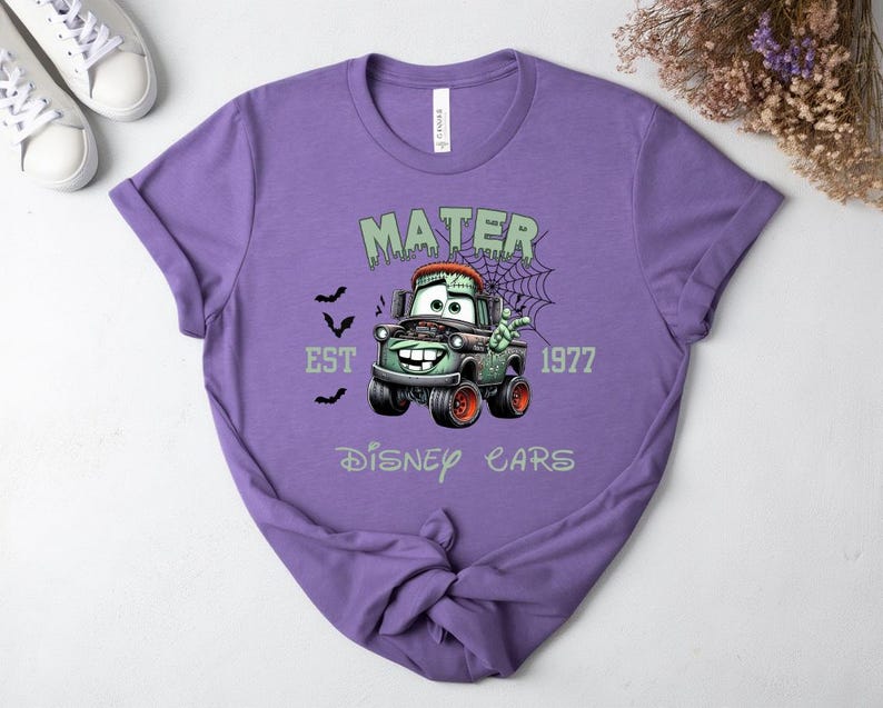 Może przedstawiać: Fioletowa koszulka z nadrukiem Mater z filmu Auta Disneya jako zombie. Projekt zawiera napisy "MATER", "EST 1977" i "Disney Cars", z nietoperzami i pajęczyną. Koszulka ma okrągły dekolt.