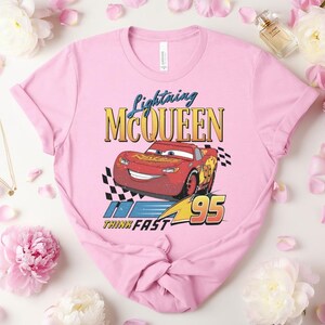 May include: Pale pink t-shirt with a graphic of Lightning McQueen from the Cars film. The design includes the text "Lightning McQueen" above a red race car with the number 95, and the words "Think Fast".