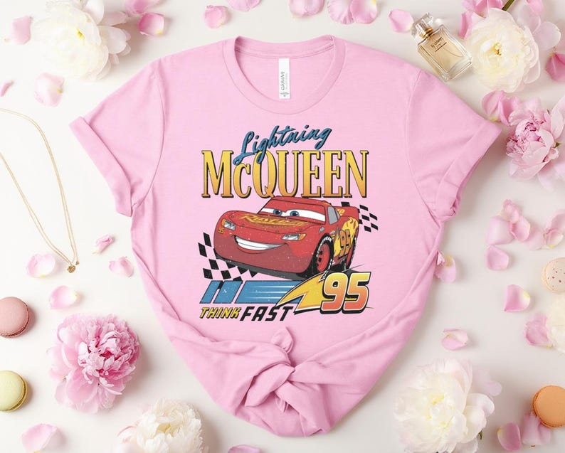 May include: Pale pink t-shirt with a graphic of Lightning McQueen from the Cars film. The design includes the text "Lightning McQueen" above a red race car with the number 95, and the words "Think Fast".