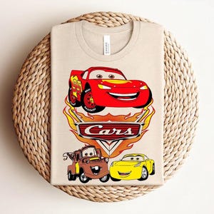 May include: Beige t-shirt featuring a colorful graphic of characters from the animated movie "Cars." The design includes a red race car, a brown tow truck, and a yellow car, with the word "Cars" in a stylized font.