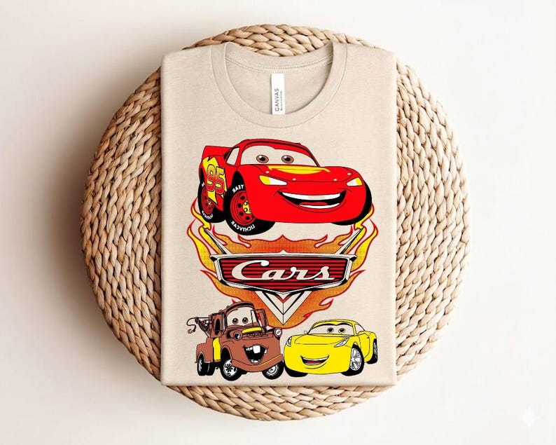 May include: Beige t-shirt featuring a colorful graphic of characters from the animated movie "Cars." The design includes a red race car, a brown tow truck, and a yellow car, with the word "Cars" in a stylized font.