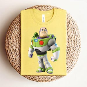 May include: Yellow t-shirt featuring a graphic of Buzz Lightyear from Toy Story. The character is depicted in his signature space ranger suit, with green, white, and purple accents. The shirt is folded and placed on a woven, circular surface.