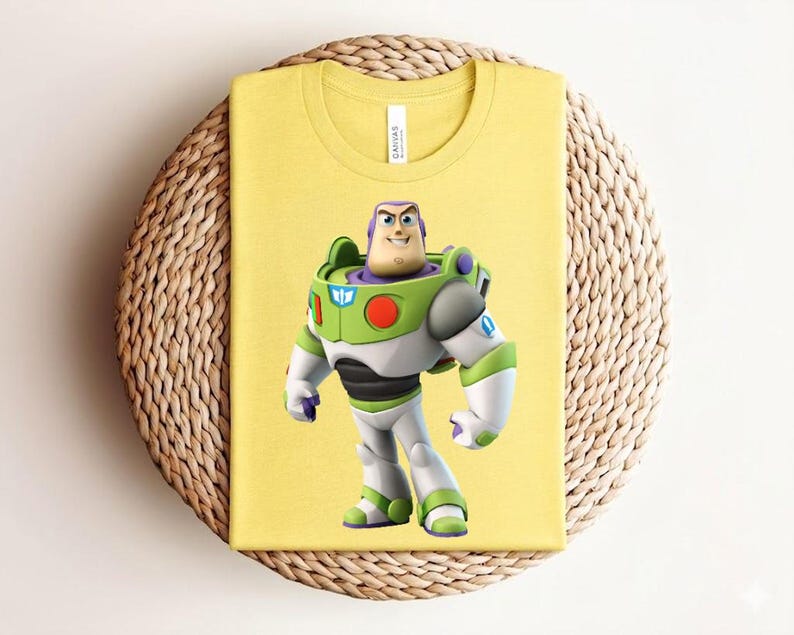 May include: Yellow t-shirt featuring a graphic of Buzz Lightyear from Toy Story. The character is depicted in his signature space ranger suit, with green, white, and purple accents. The shirt is folded and placed on a woven, circular surface.