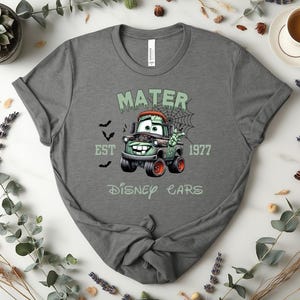 Może przedstawiać: Szara koszulka z halloweenowym motywem Złomka z filmu Auta Disneya. Projekt zawiera napis "MATER EST 1977 DISNEY CARS" z nietoperzami i pajęczyną. Koszulka ma okrągły dekolt.