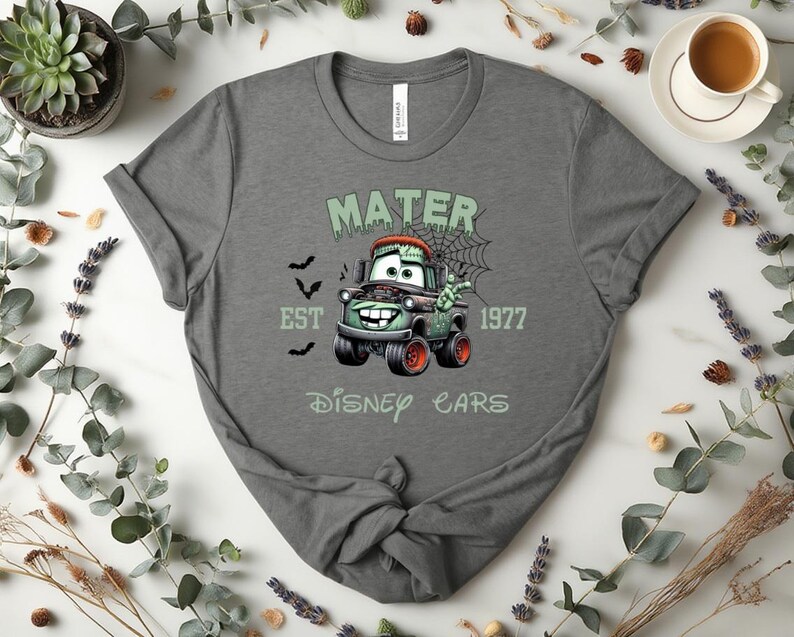 Może przedstawiać: Szara koszulka z halloweenowym motywem Złomka z filmu Auta Disneya. Projekt zawiera napis "MATER EST 1977 DISNEY CARS" z nietoperzami i pajęczyną. Koszulka ma okrągły dekolt.