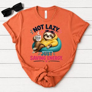 May include: Orange t-shirt featuring a sloth in sunglasses and a yellow hoodie, holding a coffee mug. The text says "NOT LAZY JUST SAVING ENERGY."