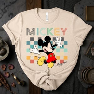 May include: Beige t-shirt featuring a colorful graphic of Mickey Mouse. The word "MICKEY" is printed across the top in various colors. Mickey Mouse is in a dynamic pose, with his arms outstretched. The shirt has a classic crew neck.